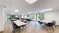 This well-appointed single-floor multipurpose facility offers a generously sized open hall designed to accommodate a variety of activities and gatherings. The main area features light-toned luxury vinyl plank flooring, a drop ceiling with recessed panel lighting, and large windows along the exterior wall that flood the space with natural light and provide lush green outdoor views. Multiple rectangular and oval tables with black chairs are arranged throughout, offering flexible seating configurations suitable for meetings, classes, or events. A wall-mounted flat-screen TV and a projection screen provide audiovisual support for presentations. One end of the hall transitions into a dedicated library nook, lined floor-to-ceiling with bookshelves and accented by warm wood slat partitioning that adds architectural interest. The space also includes a compact but fully equipped kitchenette, outfitted with white shaker-style cabinetry, light quartz countertops, a stainless steel sink, a gas range, and small appliances. Additional rooms are accessible via classic paneled doors along the rear wall, offering supplementary private spaces. The overall ambiance is clean, bright, and functional, making this an ideal setting for community programs, educational use, or organizational gatherings.