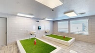 This thoughtfully designed indoor pet amenity space offers a clean, functional environment for pet relief and grooming. The room features multiple raised platforms covered in vibrant artificial turf, each accented with a decorative red fire hydrant, providing clearly defined areas for pets to use comfortably year-round regardless of outdoor conditions.

The walls are clad in large-format white subway tile to the mid-point, transitioning to a smooth painted finish above — a practical and attractive combination that ensures easy maintenance. Wood-look luxury vinyl plank flooring runs throughout the perimeter, adding warmth to the otherwise utilitarian space.

Each turf station is equipped with a retractable hose and spray attachment mounted to the wall, making cleanup straightforward and efficient. Dispensing stations mounted on the walls supply waste bags and related supplies, keeping everything pet owners need close at hand.

Overhead, flush-mounted LED panel lighting ensures the space is consistently and evenly illuminated. A textured ceiling and a security camera contribute to the room's well-managed environment. Natural light filters in through blinded windows along one wall, adding brightness to the interior.

Access is provided through a set of glass-panel double doors, offering visibility into the space from adjacent areas. The overall design balances practicality with a polished aesthetic, making it a standout amenity for pet-friendly living.