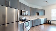 This single-floor residence showcases a thoughtfully designed interior with a consistent modern aesthetic throughout. The kitchen is a standout feature, outfitted with grey shaker-style cabinetry extending to the ceiling for ample storage, white quartz countertops, a neutral stone tile backsplash, and a full suite of stainless steel appliances including a refrigerator, electric range, over-the-range microwave, and dishwasher. A built-in desk nook adjacent to the kitchen adds a practical workspace element. Warm hardwood-style flooring flows continuously through the main living areas, creating a cohesive and inviting atmosphere. The bedroom is well-lit by a trio of large windows fitted with white horizontal blinds, allowing generous natural light to fill the room. The bathroom is finished with light large-format tile flooring, a tiled tub-shower surround accented by a decorative mosaic border, a white soaking tub, and a grey vanity with a white countertop and framed mirror. A secondary bathroom area features a separate vanity and a tiled walk-in shower, offering additional convenience. The overall design is clean, contemporary, and functional, with neutral tones and quality finishes that create a polished, move-in-ready environment.