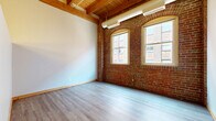 Located at 1300 West 9th Street in Cleveland, OH, this one-bedroom, one-bathroom loft-style unit showcases the character of a thoughtfully converted historic building. The living area is defined by striking exposed red brick walls, arched windows that flood the space with natural light, and warm timber ceiling beams that run the length of the room — all complemented by smooth hardwood-style flooring. The open layout creates a sense of spaciousness well beyond the unit's modest footprint.

The kitchen features hardwood-style flooring, stainless steel appliances including a full-size refrigerator, and an interior window that looks into the dramatic exposed-beam ceiling space above, adding an architectural focal point unique to the building's industrial heritage.

A carpeted hallway with continued exposed beam detailing leads to the private bedroom, a well-proportioned room that comfortably accommodates standard furnishings. The full bathroom is clean and functional, finished with white tile flooring, a combination tub and shower, a single vanity with a large mirror, and ample storage.

Throughout the unit, the interplay of original industrial elements — exposed brick, raw timber, and ductwork — with modern finishes creates a distinctive urban loft ambiance rooted in Cleveland's architectural history.