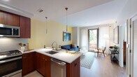 This well-appointed single-floor residence offers a thoughtfully designed open-concept layout that seamlessly connects the kitchen, dining, and living areas. The kitchen is a standout feature, equipped with rich warm-toned wood cabinetry, stainless steel appliances including a gas range, built-in microwave, refrigerator, and dishwasher, all complemented by white quartz countertops and a subway tile backsplash. A peninsula with bar seating and pendant lighting serves as both a functional workspace and a casual dining spot. The living area is bathed in natural light, with soft yellow accent walls adding warmth and vibrancy. A sliding glass door opens directly to a private outdoor balcony, offering pleasant views of surrounding greenery. Light hardwood flooring runs throughout the main living spaces, contributing to the unit's airy, modern feel. The bedroom is a comfortable retreat, featuring a large window framing lush treetop views, a bold green accent wall, and a dedicated walk-in closet providing ample storage. The bathroom is clean and contemporary, with warm wood cabinetry, a grey quartz vanity countertop, an undermount sink, and neutral tile flooring. An entry hallway with a louvered closet door completes the functional layout of this stylish residence.