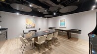 This contemporary entertainment lounge is a thoughtfully designed, single-level space that seamlessly blends social gathering and recreational amenities. The room is anchored by a large, dark stone-topped communal table surrounded by cushioned cream chairs, ideal for group activities such as board games — a chess set is prominently displayed on the table surface. Light hardwood flooring runs throughout, offering a warm contrast to the dramatic black exposed ceiling, which features visible ductwork, track lighting, and a striking array of large circular acoustic panels that add both visual interest and sound management. Along one wall, a sleek dark cabinetry wet bar with a stone countertop, integrated sink, and open shelving provides a functional refreshment station. Entertainment options abound: a full-length shuffleboard table occupies one end of the room, while a vibrant, comic-inspired arcade machine adds a playful focal point near the entrance. Curated framed artwork adorns the white walls, contributing to the polished yet energetic atmosphere. Recessed and track lighting ensures the space is well-lit throughout. The overall ambiance is modern and inviting, purpose-built to encourage leisure, social interaction, and entertainment.