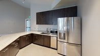 This single-floor space presents an open, versatile layout designed for communal or event use. The most striking architectural feature is the soaring vaulted ceiling, which lends a sense of grandeur and airiness throughout the main area. Exposed industrial-style ductwork runs diagonally across the ceiling, adding a contemporary design element that contrasts with the otherwise clean, neutral interior palette.

The expansive main room is finished with a patterned, herringbone-style carpet in warm beige and tan tones, providing a welcoming underfoot texture across the generous floor area. Recessed lighting is distributed throughout the ceiling, ensuring even illumination across the space. One wall features a large flat-screen television flanked by framed coastal artwork, with windows on either side allowing natural light to filter in. Double glass doors provide access to an adjacent area.

A centrally positioned kitchen area serves as a functional hub within the space. It features dark-toned cabinetry extending to the ceiling, light-toned stone countertops, and stainless steel appliances including a French door refrigerator, microwave, and dishwasher. A peninsula-style counter provides additional workspace or serving surface. Additional framed artwork and a separate door lead to what appears to be a secondary room, rounding out this well-appointed, multi-functional space.