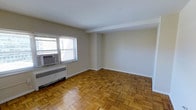 This single-floor space showcases a timeless interior defined by warm, patterned parquet hardwood flooring that adds character and visual depth throughout the main living area. The neutral, light-toned walls create a bright and versatile backdrop, lending the space a clean and open feel. Natural light filters in through a window fitted with horizontal blinds and a wall-mounted air conditioning unit, ensuring both comfort and illumination throughout the day. A baseboard radiator runs beneath the window, providing reliable in-unit heating during cooler months. The open layout of the main living area offers flexibility for a variety of furniture arrangements, making it adaptable to different uses and lifestyles. The smooth white ceiling and simple architectural lines contribute to the space's understated, classic aesthetic. Subtle structural details, including a subtle soffit element near the room's corner, add a layer of architectural interest without disrupting the overall flow. Overall, the space presents a well-maintained, functional interior with enduring design elements that balance practicality with a warm, welcoming ambiance.