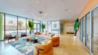 This single-floor amenity space offers a generously proportioned interior defined by bold, contemporary design and a lively, curated aesthetic. The entrance welcomes visitors through floor-to-ceiling glass walls that flood the space with natural light and provide open street views, anchored by large tropical plants that add a lush, resort-like quality. Sculptural ring-shaped pendant lighting hangs above a central lounge furnished with plush neutral sofas, vibrant tropical-print area rugs, and rounded cognac leather armchairs — creating a warm yet stylish gathering area. A pool table and additional seating nooks are positioned nearby, alongside a large-screen TV and communal table, making the space as functional as it is visually striking. The hallway transitions through a gallery-like corridor featuring framed contemporary artwork, bold orange accent walls, and a colorful mosaic tile wainscoting that adds a playful, artistic energy. A dedicated office area and secondary lounge room feature a dramatic botanical-patterned feature wall in deep green paired with a fluted column and plush carpeting, offering a more intimate retreat. Throughout, polished light stone flooring, teal panel molding details, and carefully selected furnishings tie the diverse design elements into a cohesive, vibrant interior.