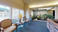 This well-appointed single-floor communal space is thoughtfully designed to offer a warm and inviting atmosphere across several distinct functional areas. The main lounge features plush seating arrangements centered around a classic white fireplace mantel, wall-mounted television, and warm ambient wall sconces, all set atop richly patterned blue carpeting that flows throughout the space. Decorative columns elegantly define transitions between areas without disrupting the open feel of the floor plan.

A bright dining nook, illuminated by a wrought-iron chandelier, offers an intimate setting with a dark wood round table, leather-upholstered chairs, and large multi-pane windows that frame lush outdoor greenery, bathing the area in natural light. Adjacent to the dining area is a functional kitchen equipped with white cabinetry, a tile backsplash, smooth countertops, and a raised bar with barstool seating — ideal for casual gatherings.

One of the most distinctive features of the space is a skylit indoor garden atrium, where an abundant collection of tropical plants and natural stone elements create a serene, nature-inspired focal point. Nearby, a sitting area with classic upholstered armchairs and large windows further enhances the connection to the outdoors. The overall ambiance is comfortable, refined, and community-oriented.