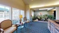 This well-appointed single-floor communal space is thoughtfully designed to offer a warm and inviting atmosphere across several distinct functional areas. The main lounge features plush seating arrangements centered around a classic white fireplace mantel, wall-mounted television, and warm ambient wall sconces, all set atop richly patterned blue carpeting that flows throughout the space. Decorative columns elegantly define transitions between areas without disrupting the open feel of the floor plan.

A bright dining nook, illuminated by a wrought-iron chandelier, offers an intimate setting with a dark wood round table, leather-upholstered chairs, and large multi-pane windows that frame lush outdoor greenery, bathing the area in natural light. Adjacent to the dining area is a functional kitchen equipped with white cabinetry, a tile backsplash, smooth countertops, and a raised bar with barstool seating — ideal for casual gatherings.

One of the most distinctive features of the space is a skylit indoor garden atrium, where an abundant collection of tropical plants and natural stone elements create a serene, nature-inspired focal point. Nearby, a sitting area with classic upholstered armchairs and large windows further enhances the connection to the outdoors. The overall ambiance is comfortable, refined, and community-oriented.