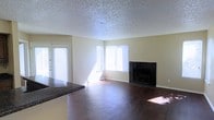 This single-floor space offers a functional and comfortable layout with a welcoming open-concept design. The living area serves as the heart of the home, showcasing rich, dark hardwood flooring that extends throughout and adds warmth to the space. A standout feature is the elegant dark-tiled fireplace, framed in sleek black tile, which serves as a natural focal point and adds a touch of sophistication to the room.

Natural light fills the living area through multiple windows, including a large window facing the exterior and a set of French doors that open to the outside, creating a bright and airy atmosphere. The neutral-toned walls complement the overall palette and provide a versatile backdrop for a variety of décor styles.

A textured popcorn ceiling adds a classic residential character to the space. The kitchen area is partially visible, featuring dark granite-style countertops that echo the tones found in the fireplace surround, creating a cohesive aesthetic flow throughout the space. The open sightline between the kitchen and living area enhances the sense of spaciousness and makes the layout ideal for everyday living and entertaining.