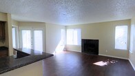 This single-floor space offers a functional and comfortable layout with a welcoming open-concept design. The living area serves as the heart of the home, showcasing rich, dark hardwood flooring that extends throughout and adds warmth to the space. A standout feature is the elegant dark-tiled fireplace, framed in sleek black tile, which serves as a natural focal point and adds a touch of sophistication to the room.

Natural light fills the living area through multiple windows, including a large window facing the exterior and a set of French doors that open to the outside, creating a bright and airy atmosphere. The neutral-toned walls complement the overall palette and provide a versatile backdrop for a variety of décor styles.

A textured popcorn ceiling adds a classic residential character to the space. The kitchen area is partially visible, featuring dark granite-style countertops that echo the tones found in the fireplace surround, creating a cohesive aesthetic flow throughout the space. The open sightline between the kitchen and living area enhances the sense of spaciousness and makes the layout ideal for everyday living and entertaining.