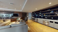 This thoughtfully designed single-floor amenity lounge offers a versatile, open-plan layout that seamlessly blends multiple functional zones. Warm hardwood flooring runs throughout, complementing the contemporary design aesthetic defined by tray ceilings with recessed lighting and a statement sputnik chandelier.

The main lounge area features generous sectional and modular sofas arranged around a central coffee table, creating inviting spaces for relaxed gatherings. A dedicated seating nook with upholstered armchairs and a side table provides a quieter retreat, while a separate cozy lounge corner includes a mustard-toned sofa and wall-mounted flat-screen TV framed by textured grey wall panels and lush indoor plants.

The wet bar area is a standout feature, showcasing stainless steel appliances, a double sink with marble-look countertops, grey cabinetry with gold hardware, and dramatic black subway tile backsplash beneath open wooden shelving. Rich wood-paneled accent walls anchor the television wall opposite.

Floor-to-ceiling sliding glass doors flood the space with natural light and provide direct access to an outdoor courtyard equipped with communal dining seating, blurring the boundary between interior comfort and outdoor living.