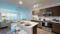 This single-floor residence showcases a contemporary design with a cohesive neutral palette and modern finishes throughout. The open-concept kitchen serves as a central feature, equipped with full-height dark wood-grain cabinetry, a white quartz countertop island with bar seating, stainless steel appliances including a range, microwave, dishwasher, and refrigerator, and a white subway tile backsplash. Wood-style plank flooring extends through the kitchen and main living areas, while recessed and track lighting ensures ample illumination.

The living room is bright and welcoming, benefiting from a large window with views of the surrounding greenery and wood-style flooring underfoot. Ceiling fans are present in both the living area and bedrooms for added comfort.

Two bedrooms feature plush carpeting, natural light from generously sized windows, and ceiling fans. The primary bedroom includes direct access to a dedicated bathroom fitted with a walk-in glass-enclosed shower, a wood-grain vanity with white quartz countertop, and marble-style tile flooring. A second bathroom mirrors this refined aesthetic, offering a soaking tub and shower combination with the same high-quality tile and vanity finishes. Overall, the space presents a clean, contemporary ambiance with thoughtful design details throughout.