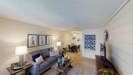 This single-floor residence offers a thoughtfully designed open-concept layout that seamlessly connects the living and dining areas, creating a cohesive and functional living environment. The living room is generously sized, featuring plush wall-to-wall carpeting that adds warmth underfoot and a textured ceiling that contributes to the room's cozy character. Neutral-toned walls provide a versatile backdrop, complemented by tasteful wall art that adds a contemporary aesthetic touch.

The living space flows naturally into the dining area, which is outfitted with a ceiling fan that promotes air circulation and comfort throughout the day. The open sightlines between the two areas make the combined space feel expansive and inviting, ideal for both everyday living and entertaining.

Decorative accents in navy blue tones are carried throughout both areas, creating a consistent and stylish color palette. A partial view of an adjacent room or entryway hints at additional functional spaces within the residence. The overall ambiance is calm and residential, with warm lighting from both ceiling fixtures and floor lamps enhancing the comfort of the interior. The layout and finishes reflect a practical yet attractive design sensibility suited for comfortable day-to-day living.