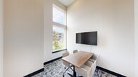 Located at 4114 FM 2920 Road in Spring, Texas, this single-room conference space delivers a modern, professional environment well-suited for small meetings and collaborative sessions. Despite its compact footprint, the room feels notably open and airy thanks to dramatically tall ceilings that draw the eye upward, amplifying the sense of vertical space.

A large, floor-to-ceiling window anchors one wall, flooding the interior with abundant natural light and offering views of the surrounding landscaped grounds. The crisp, white walls complement the bright atmosphere, while dark baseboards provide a sharp, contemporary contrast that defines the room's clean architectural lines.

At the center of the space sits a rectangular wood-topped conference table surrounded by four upholstered chairs in a neutral tone, offering comfortable seating for small group gatherings. A wall-mounted flat-screen display is positioned directly across from the window, making it ideal for presentations or video conferencing.

The floor is finished with a patterned carpet featuring a geometric, multi-tonal design that adds visual interest without overwhelming the minimalist aesthetic. A glass-paneled door with a dark frame provides both privacy and transparency, maintaining a sense of openness. Overall, the space strikes a balance between functionality and modern design.