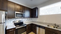 This two-story residence showcases a well-appointed kitchen as a central highlight of the main floor. The kitchen is designed with rich, dark espresso-toned cabinetry that extends floor to ceiling, providing generous storage space. Granite countertops in a light, speckled finish offer ample prep surface and contrast elegantly with the dark cabinetry. A full suite of stainless steel appliances is included, featuring a refrigerator, electric range, built-in microwave, and dishwasher, delivering a cohesive, modern aesthetic. A mosaic glass tile backsplash in neutral tones adds visual texture between the upper and lower cabinets. Light hardwood flooring runs throughout the open main level, contributing to a bright and airy atmosphere. A window above the sink area allows natural light to filter in, enhancing the overall feel of the space. The kitchen opens toward the living area, where a staircase leads to the second floor, indicating a practical, open-concept layout. The clean lines, neutral wall tones, and consistent finishes throughout create a polished, contemporary environment suited for everyday living.