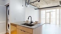Located at 2200 Market Street in Denver, CO, this studio offers a thoughtfully designed open-concept layout that blends industrial aesthetics with modern finishes. The living area features warm hardwood flooring and an exposed concrete ceiling with visible ductwork, steel beams, and track lighting — a signature loft-style look that lends the space a distinctly urban character. French doors open onto a private balcony, flooding the interior with natural light and providing views of the surrounding Denver cityscape.

The kitchen is a functional centerpiece, anchored by a peninsula island with a sleek gray countertop, undermount sink, and matte black faucet. Light wood cabinetry, white subway tile backsplash, and stainless steel appliances — including a refrigerator, range, and over-the-range microwave — complete the well-appointed cooking space. A convenient in-unit washer and dryer are tucked neatly behind louvered closet doors adjacent to the kitchen.

The bathroom continues the modern design language, featuring light wood cabinetry, a white undermount sink, gray countertop, matte black hardware, a large mirror, and hardwood flooring. Clean white walls throughout the studio enhance the sense of openness and light, making this compact space feel airy and inviting.