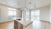 Located at 4501 E Warren Ave in Denver, Colorado, this modern single-floor residence offers a thoughtfully designed open-concept layout filled with natural light. The kitchen serves as the heart of the home, featuring a large center island with a built-in sink and quartz countertops, two globe pendant lights, two-tone cabinetry in slate blue and white, a white subway tile backsplash, and stainless steel appliances including a range and microwave. Light hardwood flooring extends throughout the main living areas, complementing the clean, neutral palette. The living area opens directly to a private balcony through floor-to-ceiling glass doors and expansive windows, offering elevated views of the surrounding Denver landscape. A ceiling fan provides additional comfort in this bright, airy space. The bedrooms are carpeted and generously proportioned, each equipped with walk-in closets featuring built-in shelving and rail systems. Two bathrooms round out the unit: one features a soaking tub with large-format wood-look tile surround, a gray vanity, and access to a walk-in closet; the second offers a glass-enclosed walk-in shower with matching tile work, a blue vanity, and hardwood-style flooring. The overall ambiance is clean, contemporary, and move-in ready.