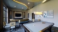 This vibrant game room is thoughtfully designed to offer both entertainment and relaxation in a single, well-appointed space. The room's most striking architectural feature is its dramatic multi-tiered ceiling, which incorporates a bold circular accent element with warm ambient uplighting, adding depth and visual interest overhead. Recessed lighting throughout ensures the space is well-lit and inviting.

At the heart of the room sits a full-size billiards table, positioned beneath a sleek rectangular pendant light that highlights it as the focal point of the space. Surrounding the table, an array of comfortable seating options are arranged throughout the room, including a curved sectional sofa upholstered in a blue and grey patterned fabric, as well as accent chairs and bistro-style tables suited for casual gathering.

A dedicated media zone features a large flat-screen television mounted within a framed wall niche flanked by decorative elements, with a low credenza below providing additional storage. Contemporary wall art and mirrored accents contribute to the modern aesthetic. The patterned carpet grounds the space with texture and color, complementing the neutral warm tones of the walls. Overall, the game room delivers a sophisticated yet comfortable atmosphere ideal for socializing and recreation.