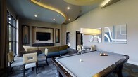 This vibrant game room is thoughtfully designed to offer both entertainment and relaxation in a single, well-appointed space. The room's most striking architectural feature is its dramatic multi-tiered ceiling, which incorporates a bold circular accent element with warm ambient uplighting, adding depth and visual interest overhead. Recessed lighting throughout ensures the space is well-lit and inviting.

At the heart of the room sits a full-size billiards table, positioned beneath a sleek rectangular pendant light that highlights it as the focal point of the space. Surrounding the table, an array of comfortable seating options are arranged throughout the room, including a curved sectional sofa upholstered in a blue and grey patterned fabric, as well as accent chairs and bistro-style tables suited for casual gathering.

A dedicated media zone features a large flat-screen television mounted within a framed wall niche flanked by decorative elements, with a low credenza below providing additional storage. Contemporary wall art and mirrored accents contribute to the modern aesthetic. The patterned carpet grounds the space with texture and color, complementing the neutral warm tones of the walls. Overall, the game room delivers a sophisticated yet comfortable atmosphere ideal for socializing and recreation.