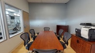 This single-floor office space is thoughtfully designed to serve both collaborative and wellness-oriented functions. The centerpiece of the space is a well-appointed conference room featuring a substantial warm-toned wood conference table surrounded by ergonomic mesh-back chairs, providing ample seating for team meetings and discussions. The room is finished with neutral gray walls, a drop tile ceiling, and light hardwood-style flooring that lends a clean, professional aesthetic. Storage is provided via rich wood cabinetry units, and a multifunction printer station is conveniently positioned along one wall. A keypad-secured door ensures controlled access to the room.

A standout architectural feature is the interior window wall that offers a direct sightline into an adjoining fitness center. This well-equipped gym area includes treadmills, an elliptical machine, and additional cardio equipment, all visible through a series of large framed windows. A wall-mounted television adds to the functionality of the fitness space. The conference room also features exterior-facing windows fitted with cellular shades, allowing for adjustable natural light and a degree of privacy. The overall design integrates a modern, functional office environment with on-site wellness amenities, reflecting a workspace built with employee productivity and well-being in mind.