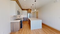 This contemporary two-story home in Phoenix, AZ offers a thoughtfully designed layout spread across two floors. The ground level houses an attached garage finished with durable epoxy-coated flooring, a glass-panel door, and direct interior access — a clean, functional space equipped with a water heater and electrical panel. A staircase leads to the main living level above, where light hardwood flooring flows throughout.

The designer kitchen is a standout feature, showcasing a striking marble-look quartz island, two-tone cabinetry combining crisp white uppers with warm wood-grain lowers, stainless steel appliances including a gas range, and elegant glass pendant lighting. The open layout and recessed lighting contribute to a bright, welcoming atmosphere.

Both bedrooms are generously proportioned, each featuring light hardwood floors, black-framed sliding windows with natural views, and ceiling fans. The spacious primary bedroom includes its own closet and an en suite bathroom outfitted with a wood-tone floating vanity, white quartz countertop, brushed gold fixtures, stone tile flooring, and a glass-enclosed walk-in shower. The secondary bathroom features similar warm finishes with a tub-shower combination and tasteful tile surround. Two closets offer ample storage throughout the home.