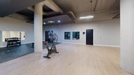Located at 1835-1849 Prospect Ave in Cleveland, OH, this fitness facility offers a thoughtfully designed, single-floor gym space that accommodates a wide range of workout needs. The open floor plan is defined by light wood flooring in the cardio and open exercise areas, transitioning to durable rubber flooring in the strength training zones — a practical and purposeful design choice.

Sturdy concrete columns throughout the space lend an industrial character, complemented by exposed ceiling infrastructure and a combination of recessed lighting and overhead fluorescent strips that ensure the space is well-illuminated throughout. The walls are painted in muted tones of sage green and light gray, creating a focused, energizing atmosphere.

The gym is equipped with a variety of fitness equipment, including stationary bikes, an elliptical trainer, a treadmill, and a cable machine, as well as a power rack with weight plates for strength training. A tiered dumbbell rack with a full range of free weights sits against a mirrored wall, enhancing the sense of depth in the space. Stability and medicine balls on a dedicated storage rack round out the functional training options.

Wall-mounted mirrors in multiple zones allow users to monitor form and contribute to the open, airy feel of the facility. Multiple entry and exit points provide easy accessibility throughout this generously sized fitness space.