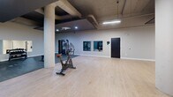 Located at 1835-1849 Prospect Ave in Cleveland, OH, this fitness facility offers a thoughtfully designed, single-floor gym space that accommodates a wide range of workout needs. The open floor plan is defined by light wood flooring in the cardio and open exercise areas, transitioning to durable rubber flooring in the strength training zones — a practical and purposeful design choice.

Sturdy concrete columns throughout the space lend an industrial character, complemented by exposed ceiling infrastructure and a combination of recessed lighting and overhead fluorescent strips that ensure the space is well-illuminated throughout. The walls are painted in muted tones of sage green and light gray, creating a focused, energizing atmosphere.

The gym is equipped with a variety of fitness equipment, including stationary bikes, an elliptical trainer, a treadmill, and a cable machine, as well as a power rack with weight plates for strength training. A tiered dumbbell rack with a full range of free weights sits against a mirrored wall, enhancing the sense of depth in the space. Stability and medicine balls on a dedicated storage rack round out the functional training options.

Wall-mounted mirrors in multiple zones allow users to monitor form and contribute to the open, airy feel of the facility. Multiple entry and exit points provide easy accessibility throughout this generously sized fitness space.