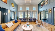 This thoughtfully designed single-floor community space makes a striking first impression with its soaring double-height lobby, wrapped in rich teal-blue paneled walls, coffered ceiling details, and an abundance of natural light streaming through tall, multi-pane windows. The welcoming lounge area features a warm caramel leather sofa, woven accent chairs, and a patterned herringbone hardwood floor that flows throughout the space, tying each area together cohesively.

A built-in workstation along the window wall provides an ideal co-working setup, with views of lush greenery beyond. Adjacent to the main lounge, a glass-enclosed private office offers a quiet, focused workspace accented by a textured chevron-patterned accent wall and modern ergonomic seating with ample natural light from a side window.

The space also includes a well-appointed coffee bar and kitchenette, featuring quartz countertops, a geometric tile backsplash, floating wood shelves, and a wall-mounted television — creating a casual gathering spot for residents or visitors. A clean, light-filled hallway with the same herringbone flooring connects the spaces and leads to a restroom. Throughout, the design balances modern aesthetics with functional amenities, resulting in an inviting and polished environment.