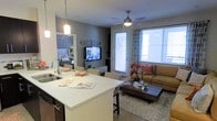 This single-floor space is designed with an open-concept layout that seamlessly connects the kitchen, dining, and living areas into one cohesive living environment. The kitchen features a large white countertop island with an undermount stainless steel sink, pendant lighting overhead, and bar-height seating — making it ideal for both meal preparation and casual dining. Dark cabinetry provides a striking contrast to the bright countertops, adding a contemporary edge to the space.

The dining area transitions naturally from the kitchen island, offering additional seating and a clear sightline into the living room. The living room is anchored by a warm-toned sofa, a dark wood coffee table atop a plush area rug, and a wall-mounted flat-screen television. A ceiling fan with an integrated light fixture adds both functionality and a finishing touch to the room.

Large windows dressed with patterned drapes and adjustable blinds allow generous natural light to fill the space, creating a bright and welcoming atmosphere. A glass-panel door provides access to an outdoor area. Soft grey walls throughout lend a neutral, modern backdrop that complements the carefully curated furnishings. A glimpse into the adjacent bedroom reveals a well-appointed sleeping area, reinforcing the cohesive design aesthetic found throughout the entire space.