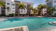 Located at 7120 Okeechobee Blvd in West Palm Beach, Florida, this outdoor amenity space showcases a generously sized resort-style swimming pool as its centerpiece. The pool features crystal-clear blue water and is surrounded by an expansive paver deck finished in a sophisticated multi-tone stone pattern, providing ample room for lounging and recreation.

Numerous cushioned chaise lounge chairs are arranged throughout the deck, offering residents a variety of sunbathing and relaxation options. A covered gazebo with a pitched roof serves as a shaded retreat within the pool area, providing shelter from the Florida sun while maintaining an open, airy feel. Additional shade is provided by green market umbrellas strategically positioned around the deck, along with striped cabana-style curtained structures.

Tall, mature royal palms and lush tropical plantings frame the entire space, reinforcing a resort-like atmosphere consistent with South Florida living. The pool area is embraced on multiple sides by contemporary multi-story residential buildings featuring modern architecture with clean lines, neutral tones, and private balconies that overlook the amenity space. The combination of abundant natural sunlight, vibrant tropical greenery, and thoughtfully arranged outdoor furnishings creates an inviting and well-appointed outdoor environment ideal for year-round enjoyment.