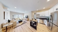 This thoughtfully designed single-floor unit offers a bright, open-concept layout that seamlessly connects the living, dining, and kitchen areas. Light hardwood flooring runs throughout, lending warmth and continuity to the space. Recessed lighting illuminates the living room, which features large windows that invite natural light and a wall-mounted mini-split system for climate control.

The kitchen is well-appointed with white shaker-style cabinetry, dark countertops, and a full suite of stainless steel appliances, including a refrigerator, range, microwave, and dishwasher. A freestanding island with a butcher block top provides additional prep space and casual seating.

The bedroom is a serene retreat, benefiting from a window with garden views and continued hardwood flooring. Two bathrooms serve the unit, both finished with striking marble-look tile flooring, white cabinetry, dark countertops, and matte black hardware accents. One bathroom includes a window for added ventilation and natural light.

A dedicated office or flex room rounds out the layout, featuring corner windows on two walls that flood the space with daylight — ideal for productive work or use as an additional bedroom. The overall palette of warm neutrals and clean finishes creates a cohesive, contemporary ambiance throughout.