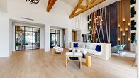 Located at 100 Boucle Jeanne Cir in Maitland, Florida, this single-floor lobby space makes an immediate impression with its soaring double-height ceilings and exposed natural wood beam trusses that span the full width of the room. Light-toned hardwood flooring runs throughout, lending warmth and continuity to the open layout.

A dramatic focal point anchors the main lounge area: an artistic installation of angled wooden rods and hanging rope lights cascades from the ceiling against a dark textured accent wall adorned with vertical greenery, creating a bold visual contrast against the crisp white walls. A built-in white sofa accented with navy and patterned throw pillows sits beneath, accompanied by sculptural wood-and-white chairs and natural log side tables.

Arched double entry doors framed by floor-to-ceiling windows invite abundant natural light into the welcoming entrance, where sheer drapery softens the generous glazing. A seating nook near the windows features striped accent chairs and elegant lantern-style candle holders. A unique live-edge wood reception console, paired with a round mirror and lush greenery, serves as a refined front desk area. Steel-framed glass partition doors offer a glimpse into an adjacent lounge beyond. The overall ambiance is sophisticated yet warm, blending organic textures with modern design.
