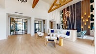 Located at 100 Boucle Jeanne Cir in Maitland, Florida, this single-floor lobby space makes an immediate impression with its soaring double-height ceilings and exposed natural wood beam trusses that span the full width of the room. Light-toned hardwood flooring runs throughout, lending warmth and continuity to the open layout.

A dramatic focal point anchors the main lounge area: an artistic installation of angled wooden rods and hanging rope lights cascades from the ceiling against a dark textured accent wall adorned with vertical greenery, creating a bold visual contrast against the crisp white walls. A built-in white sofa accented with navy and patterned throw pillows sits beneath, accompanied by sculptural wood-and-white chairs and natural log side tables.

Arched double entry doors framed by floor-to-ceiling windows invite abundant natural light into the welcoming entrance, where sheer drapery softens the generous glazing. A seating nook near the windows features striped accent chairs and elegant lantern-style candle holders. A unique live-edge wood reception console, paired with a round mirror and lush greenery, serves as a refined front desk area. Steel-framed glass partition doors offer a glimpse into an adjacent lounge beyond. The overall ambiance is sophisticated yet warm, blending organic textures with modern design.
