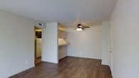 This single-floor space presents a clean, functional layout designed for comfortable everyday living. The main living area is finished with warm, dark wood-style hardwood flooring that extends throughout, lending a cohesive and grounded feel to the interior. Neutral-toned walls and white baseboards keep the space feeling bright and well-maintained.

A ceiling fan with an integrated light fixture is centrally positioned overhead, providing both air circulation and ambient lighting to the living area. A standout architectural detail is a built-in desk or counter nook tucked into the wall, offering a dedicated surface for work or display without occupying additional floor space.

An open passageway connects the living area to the kitchen, which is partially visible and features dark cabinetry and a white refrigerator, suggesting a compact but functional cooking space. A ceiling vent ensures proper climate control throughout. The overall ambiance is understated and move-in ready, with simple finishes that provide a neutral canvas adaptable to a variety of furnishing styles and personal aesthetics.