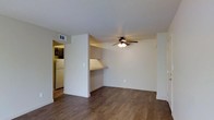 This single-floor space presents a clean, functional layout designed for comfortable everyday living. The main living area is finished with warm, dark wood-style hardwood flooring that extends throughout, lending a cohesive and grounded feel to the interior. Neutral-toned walls and white baseboards keep the space feeling bright and well-maintained.

A ceiling fan with an integrated light fixture is centrally positioned overhead, providing both air circulation and ambient lighting to the living area. A standout architectural detail is a built-in desk or counter nook tucked into the wall, offering a dedicated surface for work or display without occupying additional floor space.

An open passageway connects the living area to the kitchen, which is partially visible and features dark cabinetry and a white refrigerator, suggesting a compact but functional cooking space. A ceiling vent ensures proper climate control throughout. The overall ambiance is understated and move-in ready, with simple finishes that provide a neutral canvas adaptable to a variety of furnishing styles and personal aesthetics.