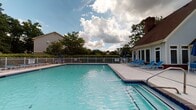 Located at 5715 Castlebridge Rd in Fredericksburg, Virginia, this single-story community pool facility offers a well-appointed outdoor recreational experience surrounded by lush greenery. The centerpiece of the property is a generously sized outdoor swimming pool with clear, blue-tinted water, framed by a wide concrete pool deck. The deck is outfitted with multiple blue lounge chairs and patio umbrellas, providing ample space for sunbathing and relaxation. A white metal fence encloses the pool area, ensuring safety while maintaining open sightlines to the mature trees and landscaping that border the property.

The interior building component is a compact, single-floor structure with a classic brick exterior and a pitched roof, directly adjacent to the pool. Inside, the entryway features white tile flooring with a welcome mat, warm wood trim wainscoting along the walls, and light blue upper walls that carry a clean, neutral aesthetic. Wood-trimmed doorways lead to restroom facilities, clearly marked with signage, as well as a carpeted interior room. A glass-panel entry door provides a direct view of the pool deck, connecting the interior and outdoor spaces seamlessly. The facility is designed to serve as a functional and comfortable amenity for the surrounding community.