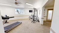 The fitness center at The Place At 2120, located at 2120 N Silverbell Rd in Tucson, Arizona, offers a well-equipped, single-floor workout facility designed to support a well-rounded exercise routine. The space is organized across several interconnected areas, each dedicated to a specific type of training.

The main workout area features neutral-toned carpet flooring throughout and bright white walls accented by a continuous beige chair rail, lending the space a clean and welcoming atmosphere. Large windows along the exterior walls invite an abundance of natural light and provide views of the surrounding palm-lined landscape, enhancing the open feel of the room. A ceiling fan provides additional ventilation and comfort.

Cardio equipment, including treadmills and a stationary bike, is positioned to take advantage of the natural light and outdoor views. A multi-station cable machine serves as the centerpiece for strength training, complemented by a dedicated free-weight area featuring a full dumbbell rack, an adjustable bench, and a wall-mounted mirror. A wall-mounted flat-screen television is also present for entertainment during workouts.

A separate alcove provides storage for a colorful set of medicine balls mounted on a wall rack, alongside an exterior door for convenient access. The overall ambiance is bright, functional, and well-organized, making it a practical fitness resource for residents.
