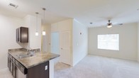 Located at 115 Amberleigh Dr in Wilmington, NC, this single-story home offers an open-concept layout that connects the kitchen and living areas seamlessly. The kitchen features dark espresso cabinetry paired with granite-style countertops and a built-in sink, complemented by stylish pendant lighting that hangs above the counter, adding a modern touch. The textured ceiling detail above the kitchen area adds architectural interest, while a ceiling fan in the living space promotes airflow and comfort. The living area is finished with plush light-colored carpeting, creating a warm and inviting atmosphere. A window allows natural light to fill the space, enhancing the bright, airy feel of the interior. Louvered closet doors provide functional storage access while maintaining a clean aesthetic. The neutral color palette throughout the home creates a versatile backdrop suitable for a variety of décor styles. Overall, the space presents a well-designed, comfortable single-floor layout ideal for everyday living in Wilmington, NC.