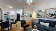 This single-floor residence offers a thoughtfully designed layout that balances comfort and functionality. The open-concept living area serves as the heart of the home, featuring warm hardwood-style flooring, vaulted ceilings with a ceiling fan, and generous natural light streaming through large windows dressed with stylish patterned drapes. The living space is inviting and well-proportioned, with room for relaxed seating arrangements and tasteful wall art that adds a contemporary touch.

The kitchen flows seamlessly from the living area via a half-wall pass-through, and is outfitted with white cabinetry accented with wood trim, granite-style countertops, and black appliances. A dedicated walk-in pantry provides ample storage, making this kitchen as practical as it is attractive.

The residence includes two bedrooms, both carpeted for comfort. The primary bedroom is bathed in natural light from multiple windows with outdoor greenery views, and features coordinated patterned drapery. The secondary bedroom also enjoys window views to the outside. The bathroom includes a full tub-shower combination with clean white tile surround.

Throughout, the space maintains a cohesive, neutral palette accented with navy and teal tones, creating a warm and welcoming ambiance well-suited for everyday living.