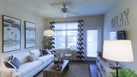 This single-floor space offers a thoughtfully designed and well-appointed interior that blends contemporary style with functional comfort. The living area serves as the focal point of the space, showcasing a cohesive design palette anchored by striking black-and-white chevron-patterned curtains that frame large windows, allowing generous natural light to fill the room. The windows are fitted with white plantation-style blinds, offering both privacy and light control. A classic ceiling fan with a brushed metal finish is centrally mounted, providing both airflow and a casual, relaxed aesthetic. The living area is furnished with a clean-lined white sofa accented with patterned throw pillows, complemented by a rustic wooden coffee table that adds warmth and contrast to the otherwise light color scheme. Dark hardwood flooring grounds the space and adds a rich, elegant touch, while a neutral area rug defines the seating arrangement. A media console along one wall provides additional storage and display space. Tasteful lighting, including a floor lamp with a drum shade, enhances the room's warm ambiance. Wall art and carefully selected décor accessories complete the space, contributing to an overall atmosphere that is both inviting and visually polished.