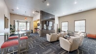 Located in Katy, TX, this single-floor community lounge space offers a well-appointed and versatile gathering area designed for both relaxation and recreation. The main living area is generously proportioned, featuring a sophisticated interior defined by patterned charcoal carpet with marble-like striping, recessed lighting, and neutral-toned walls accented by colorful abstract artwork.

The lounge area is anchored by a prominent tile-clad feature wall with a mounted flat-screen television, creating a natural focal point for seating. Cream-colored leather club chairs with built-in side tables are arranged throughout the space, offering multiple intimate seating clusters. An orange sofa along one wall adds a bold accent to the contemporary palette.

A dedicated game zone includes a full-sized pool table, while a business center area is equipped with desktop computers, cabinetry, and counter workspace — ideal for productivity. Multiple windows with horizontal blinds line the walls, allowing ample natural light to fill the room while maintaining privacy.

The hallway connects the main areas seamlessly, contributing to an efficient and open floor plan. Potted greenery and carefully chosen décor add warmth and life to the space. Overall, this community room delivers a polished, modern ambiance suited for socializing, entertainment, and everyday use.