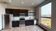 Located at 449 1/2 Bridge St NW in Grand Rapids, Michigan, this single-floor unit blends contemporary design with industrial-inspired details throughout. Light wood-style flooring runs continuously through the space, complementing the neutral gray walls and dark trim accents.

The kitchen serves as a central highlight, outfitted with dark espresso cabinetry that extends to the ceiling for ample storage, paired with light gray countertops and a crisp white subway tile backsplash. A full suite of stainless steel appliances includes a refrigerator, electric range, built-in microwave, and dishwasher. Industrial-style pipe shelving mounted on the wall adds a distinctive urban character, while a large window frames views of the surrounding Grand Rapids neighborhood and fills the space with natural light.

The bedroom features exposed ductwork overhead, lending an industrial loft aesthetic, alongside a tall window that offers elevated views of the nearby rooftops and greenery.

The bathroom is thoughtfully appointed with a dark wood vanity, gray countertop, undermount sink, and matte black fixtures. A caged wall sconce adds an industrial touch above the mirror, while a full-size tub and shower complete the room. Light wood-style flooring ties the bathroom seamlessly to the rest of the unit.