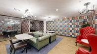 Located at 40 Locke St in Haverhill, Massachusetts, this single-floor space offers a distinctive and visually striking interior defined by its bold, colorful wallcoverings that wrap nearly every surface in a vivid pop-art-inspired pattern of teal, orange, red, and green. The main lounge area serves as the centerpiece, anchored by a pair of tufted green leather Chesterfield sofas arranged on a blue area rug atop warm hardwood flooring. A statement red tufted wingback chair adds a vibrant accent. Overhead, industrial-style pendant chandeliers with exposed globe bulbs provide stylish ambient lighting, complemented by recessed ceiling lights throughout. Multiple round marble-top tables with dark bistro-style chairs are distributed across the space, offering flexible seating arrangements for both casual gathering and small meetings. A sculptural white column near the entrance adds an architectural focal point. Glass-panel partitions with black metal frames divide the space into distinct zones, including what appears to be a private office or meeting room equipped with task chairs and a conference table, accessible via sliding glass doors. A large decorative mirror framed in an ornate dark surround adds depth to the lounge area. The overall ambiance is energetic and creative, combining eclectic design elements with functional, multi-use areas suited for collaborative or social settings.