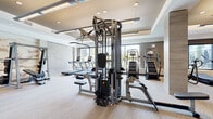 This expansive fitness center is thoughtfully designed to support a full spectrum of workout routines in a sleek, contemporary environment. The space is laid out across a single open floor plan, with distinct zones dedicated to cardio, strength training, and free weights, allowing for an efficient and comfortable workout experience.

The cardio area features multiple treadmills, stair climbers, stationary bikes, and rowing machines, all positioned along large floor-to-ceiling windows that frame views of an outdoor pool area and surrounding greenery. Natural light floods the space, creating an energizing and open atmosphere.

The strength training zone includes a multi-station cable machine, a Smith machine with an adjustable bench, and an extensive rack of dumbbells, catering to a wide range of exercises and fitness levels. Full-length mirrors are strategically placed throughout to support proper form and enhance the sense of depth and scale within the room.

Architectural details elevate the overall design, including tray ceilings with warm gold accents, recessed and pendant lighting, and textured stone-look wall panels. A decorative mural adds an artistic focal point to the weight training area. Carpeted flooring in a neutral, light-toned pattern ties the space together, while glass-paneled walls near the entrance provide transparency and connectivity to adjacent amenity areas. The overall ambiance is polished, modern, and well-appointed.