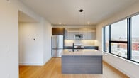 Located in Philadelphia's vibrant North Howard Street corridor, this contemporary 2-bedroom, 1-bathroom unit offers a thoughtfully designed single-floor layout with modern finishes throughout. The open-concept kitchen serves as the heart of the space, showcasing two-tone cabinetry in warm wood and cool gray, stainless steel appliances including a full-size refrigerator, electric range, and built-in microwave, as well as a large central island with a white quartz countertop and an undermount sink. Pendant lighting and under-cabinet illumination add to the kitchen's polished aesthetic, while large floor-to-ceiling windows flood the area with natural light and frame views of the surrounding Philadelphia cityscape. Light hardwood flooring flows through the main living areas, complementing the bright, neutral color palette. Both bedrooms feature plush carpeting, recessed lighting, and generous windows that bring in additional natural light and urban views. The primary bedroom includes an open closet with built-in shelving. The bathroom is sleek and modern, appointed with large-format gray tile, a glass-enclosed walk-in shower, a wall-mounted vanity with a backlit mirror, and matte black fixtures throughout. Hallways connect the spaces efficiently, with additional built-in storage closets adding practical functionality to this well-appointed urban residence.