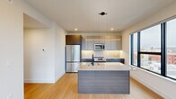 Located in Philadelphia's vibrant North Howard Street corridor, this contemporary 2-bedroom, 1-bathroom unit offers a thoughtfully designed single-floor layout with modern finishes throughout. The open-concept kitchen serves as the heart of the space, showcasing two-tone cabinetry in warm wood and cool gray, stainless steel appliances including a full-size refrigerator, electric range, and built-in microwave, as well as a large central island with a white quartz countertop and an undermount sink. Pendant lighting and under-cabinet illumination add to the kitchen's polished aesthetic, while large floor-to-ceiling windows flood the area with natural light and frame views of the surrounding Philadelphia cityscape. Light hardwood flooring flows through the main living areas, complementing the bright, neutral color palette. Both bedrooms feature plush carpeting, recessed lighting, and generous windows that bring in additional natural light and urban views. The primary bedroom includes an open closet with built-in shelving. The bathroom is sleek and modern, appointed with large-format gray tile, a glass-enclosed walk-in shower, a wall-mounted vanity with a backlit mirror, and matte black fixtures throughout. Hallways connect the spaces efficiently, with additional built-in storage closets adding practical functionality to this well-appointed urban residence.