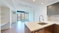 Located in the heart of downtown San Diego's East Village neighborhood, this modern high-rise unit offers a sophisticated urban living experience with sweeping city skyline views. The open-concept layout is anchored by a sleek kitchen featuring rich wood-toned cabinetry, a tile backsplash, stainless steel appliances including a French door refrigerator, microwave, and range, along with a generous island topped with quartz countertops and an integrated sink with a professional-style faucet. Light wood flooring flows throughout, complementing the clean white walls and recessed lighting. The living area is bathed in natural light from expansive floor-to-ceiling windows framed in bold black trim, with a balcony door opening to panoramic views of the San Diego skyline. The bedroom continues the floor-to-ceiling window theme, offering stunning elevated cityscape views and a ceiling fan. Built-in shelving adds functional character to the secondary room. A spacious walk-in closet with adjustable shelving and rail systems provides excellent storage. The primary bathroom is well-appointed with a dual-sink vanity, large mirror, wood cabinetry, a glass-enclosed marble-tiled shower, and direct access to the walk-in closet. Track lighting and recessed fixtures illuminate the space throughout.