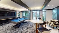 Located at 1314 Montrose Blvd in Houston's Montrose neighborhood, this contemporary game room and social lounge is designed for entertainment and community gathering. The space features a bold, modern aesthetic defined by tray ceilings with recessed lighting, geometric patterned carpeting, and a striking color palette of deep navy, teal, and white.

At the heart of the room sits a full-size pool table with a vivid blue felt surface, complemented by a wooden shuffleboard table and a classic Pac-Man arcade cabinet — offering a diverse range of recreational activities. A large flat-screen television is mounted on a dark accent wall, framed by an arrangement of decorative geometric mirrors that add visual depth to the space.

Curated artwork, including a bold graffiti-style canvas and framed prints, lends an energetic, urban character to the interior. High-top tables with modern bar seating provide casual gathering spots throughout the room, while additional lounge seating is available near floor-to-ceiling glass doors that open directly to an outdoor terrace. This seamless indoor-outdoor connection fills the space with natural light and views of the surrounding area. The overall ambiance balances a lively, social atmosphere with polished, upscale design.