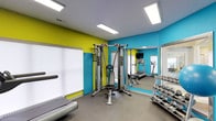 This fitness center offers a well-rounded exercise environment designed to accommodate a variety of workout routines. The space is immediately distinguished by its bold, energetic color scheme — vivid turquoise and yellow-green accent walls that create a motivating atmosphere. Large, flush-mounted ceiling lights provide bright, even illumination throughout, ensuring excellent visibility across all areas of the room.

The facility is thoughtfully equipped with a range of cardio machines, including treadmalls, positioned to make efficient use of the space. A multi-function cable machine serves as a centerpiece for strength training, complemented by a workout bench ideal for a variety of exercises. A dedicated free weights area features a full dumbbell rack, offering options for users of all fitness levels.

Strategically placed mirrors along the walls enhance the sense of openness while allowing users to monitor their form during workouts. A mounted flat-screen television adds a touch of convenience for those looking to stay entertained during cardio sessions. The flooring is finished in a durable, dark rubberized material suited for high-traffic athletic use. Overall, this fitness center combines functionality with a dynamic, welcoming design.