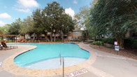 Located in Houston, TX, this single-story outdoor community space offers a well-designed resort-style environment characterized by thoughtful landscaping and recreational amenities. The centerpiece of the property is a generously sized, freeform swimming pool with clear turquoise water, complemented by a separate spa featuring a stone surround and bubbling fountain. The pool deck is finished in neutral-toned concrete and stone pavers, providing ample space for lounging with wrought-iron chaise lounges, accent tables, and umbrella-shaded dining sets arranged throughout the area.

Mature oak and crape myrtle trees provide natural shade across the grounds, creating a canopied atmosphere that enhances the outdoor experience. A covered pergola structure adds architectural interest to the pool surround. The community building features a traditional exterior with stone and brick detailing, covered porches, large windows, and stone-stepped entryways bordered by ornamental plantings including sago palms and trimmed hedges.

Access to the property is controlled through a decorative wrought-iron gate flanked by landscaped beds, ensuring a secure and welcoming entry. The overall ambiance is serene and resort-like, well-suited to the warm Houston climate.