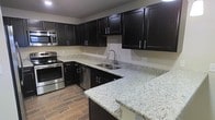 This two-story home offers a well-designed layout with thoughtfully appointed finishes throughout. The kitchen serves as a central highlight, showcasing rich dark cabinetry with sleek hardware, light granite countertops, and a full suite of stainless steel appliances including a range, microwave, and dishwasher. A built-in breakfast bar extends the counter space and provides a natural transition into the adjacent living area, which features warm, neutral carpeting and tile-look flooring. Pendant lighting above the bar adds a refined touch to the open-concept design. The kitchen's functional L-shaped layout maximizes storage and workspace, with ample upper and lower cabinetry lining the walls.

Upstairs, a generously sized room with vaulted ceilings creates an airy, open atmosphere. Dual windows allow natural light to fill the space, and a glass-paneled door provides additional brightness. The soft carpeting and angular ceiling lines give the room a welcoming, comfortable character. Neutral tones are carried throughout the home, creating a cohesive and move-in-ready aesthetic. This space combines practical functionality with modern design elements, making it a well-rounded and comfortable home across both levels.